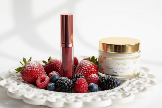 create a image of a clear lip gloss tube filled with a blend of chocolate and strawberry mixed to make the perfect color called Mixed Berry, with a pink metalic top on the tube sitting on pretty dish surrouded my mixed berries with powdered sugar spinked over them and a glass clear jar with white body butter and a gold shiny lid beside the dish and the body butter jar should read Manuki Pistachio & Magnoiia Body Butter